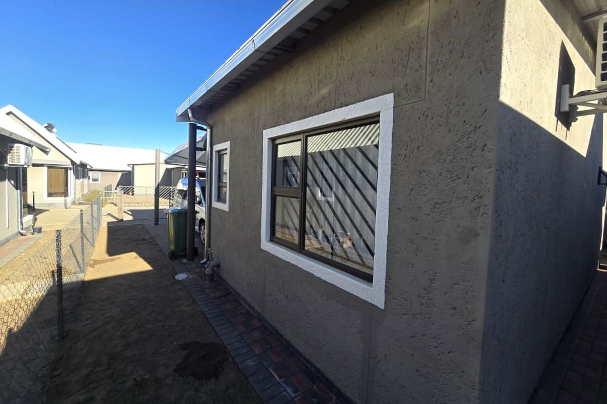 A view of the rear exterior of the apartment, featuring a textured plaster finish, aluminium windows with security bars, and a paved walkway.