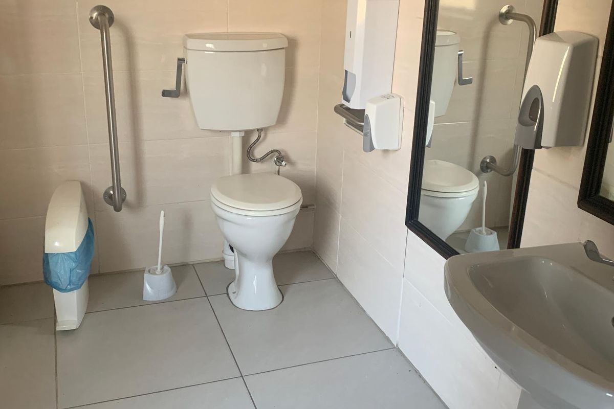 A functional office restroom featuring a toilet, pedestal sink, wall-mounted grab rails, and tiled surfaces.