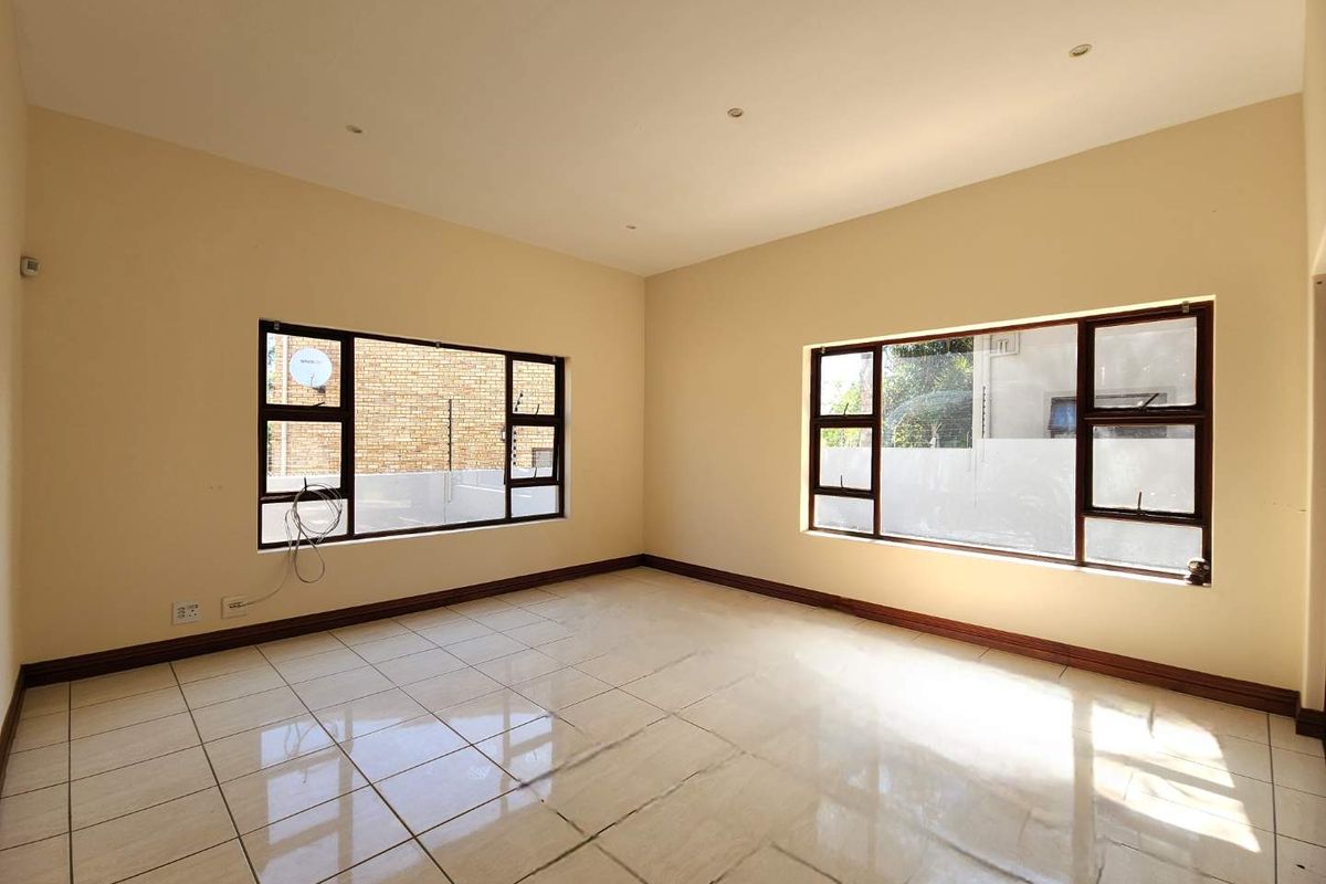 A bright, empty living room featuring large windows with dark frames and neutral ceramic floor tiles throughout.