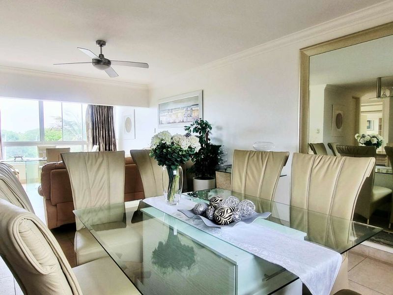 An open-plan living and dining area featuring tiled flooring, a ceiling fan, and a large wall mirror, with access to a balcony through sliding doors.