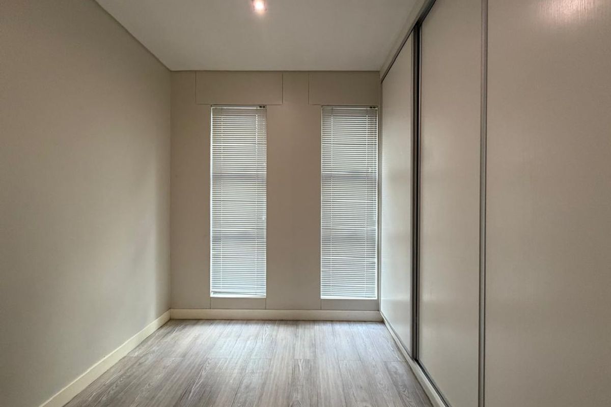 A bright, empty bedroom featuring modern wood-look flooring, built-in sliding door wardrobes, and dual windows with fitted blinds.