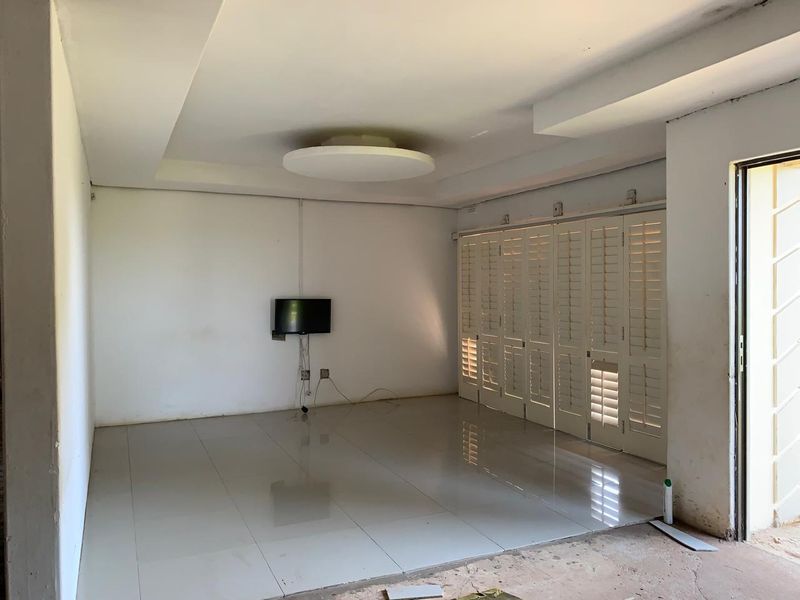 An empty living room featuring white tiled flooring, a recessed ceiling with a central light fixture, and white plantation shutters.