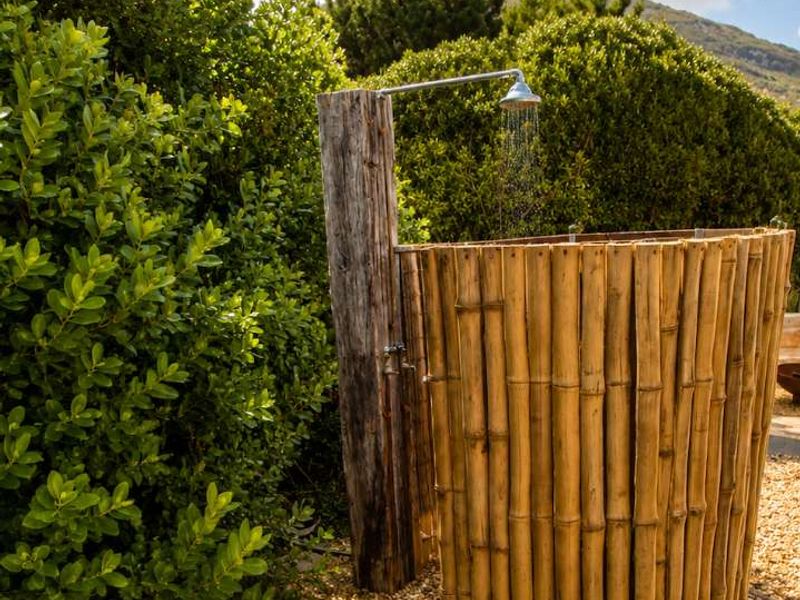 An outdoor shower area featuring a rustic timber post, shower head, and bamboo privacy screen set within a landscaped garden with gravel ground cover.