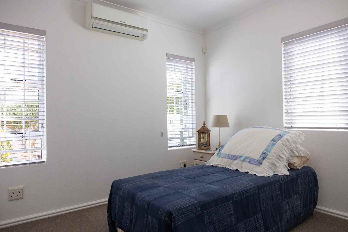 A functional bedroom featuring neutral walls, carpet flooring, multiple windows with venetian blinds, and a wall-mounted split-system air conditioning unit.