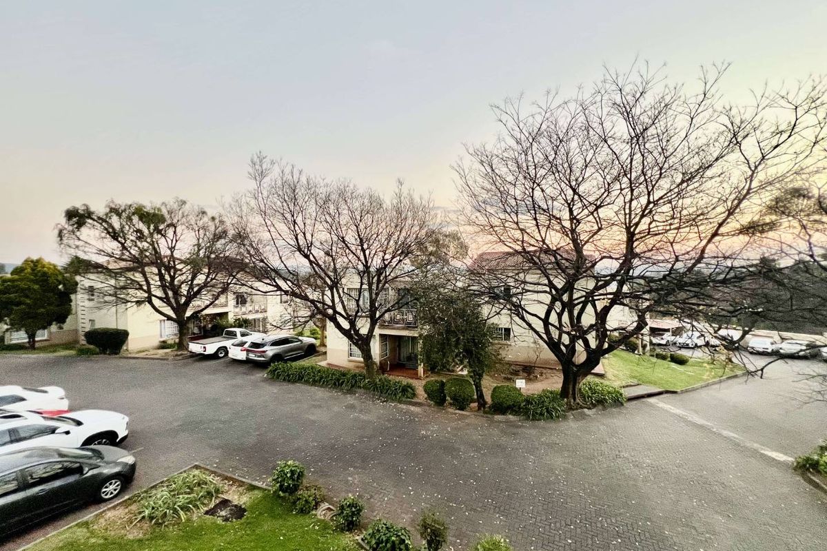 An elevated view of the apartment complex grounds, featuring paved parking areas, landscaped garden beds, and mature trees.
