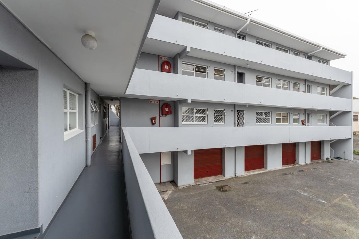 Exterior view of an apartment complex featuring concrete walkways, security bars on windows, fire safety equipment, and ground-level garage access.