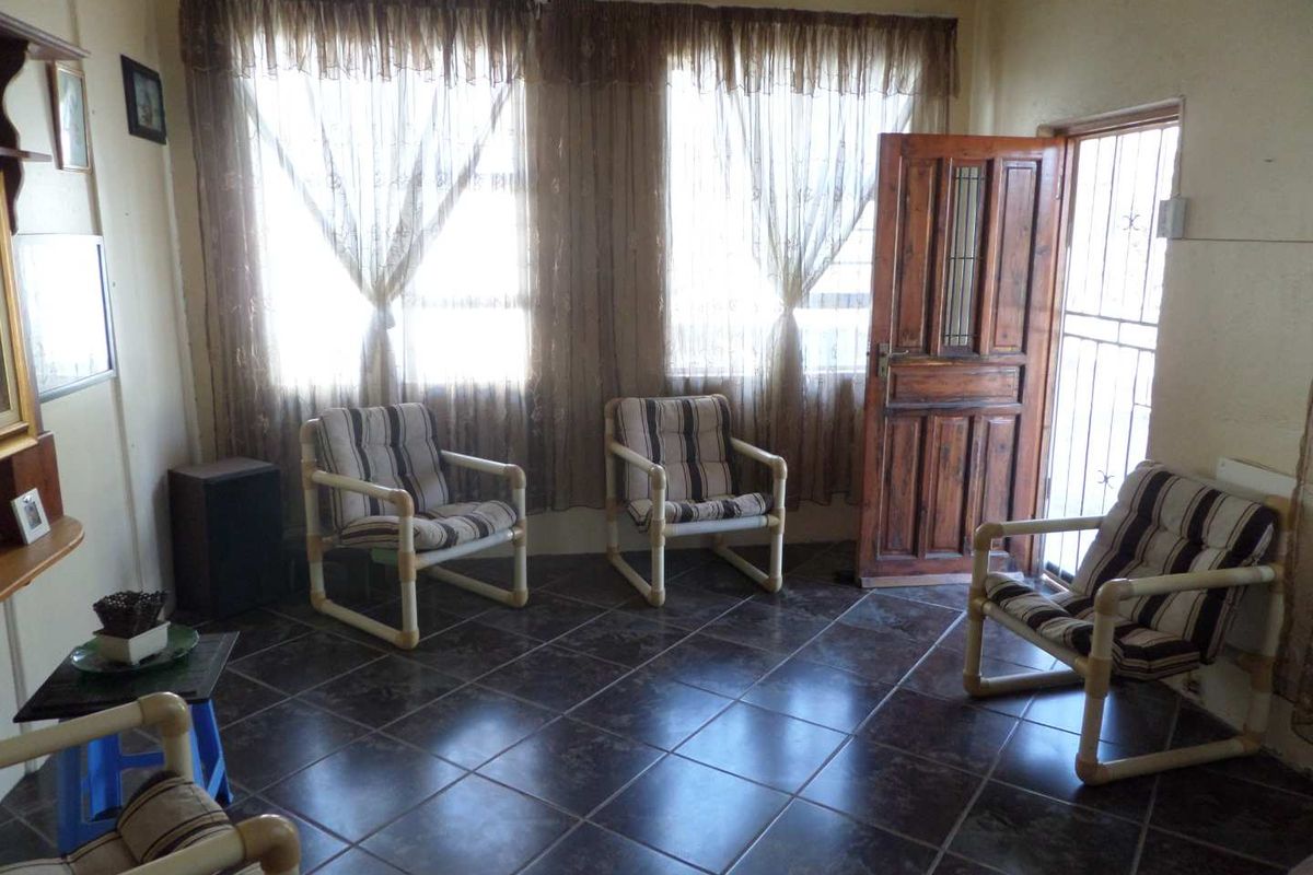 A living room featuring dark tiled flooring, large windows with curtains, and a wooden entrance door with security gate.