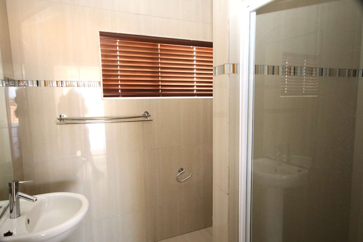 A modern bathroom featuring neutral wall tiles, a pedestal basin, a glass-enclosed shower, and a window with fitted blinds.