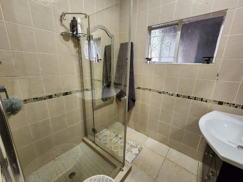 A functional bathroom featuring a glass-enclosed shower, neutral ceramic tiling with a mosaic accent border, and a pedestal washbasin.