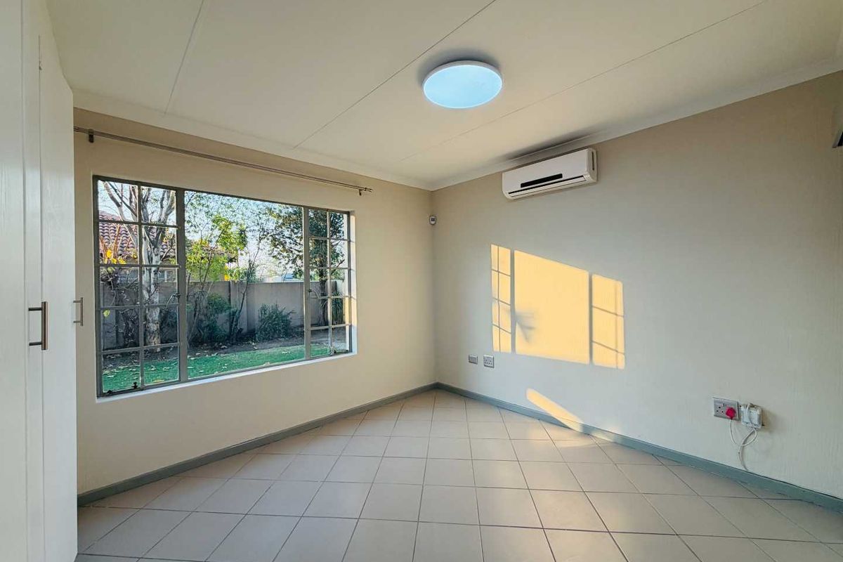 A bright, empty bedroom featuring tiled flooring, a built-in cupboard, and a wall-mounted air conditioning unit with a view of the garden.