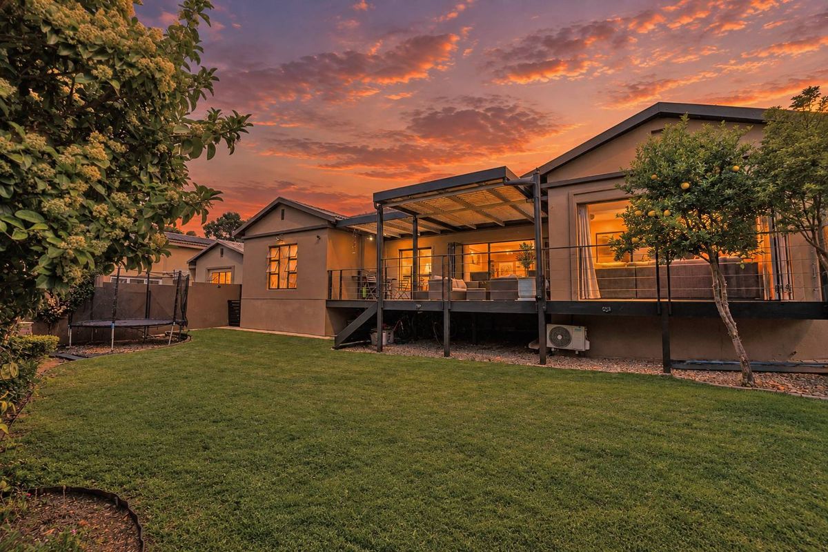 Rear view of the property featuring a raised timber deck with a pergola, overlooking a well-maintained garden at sunset.
