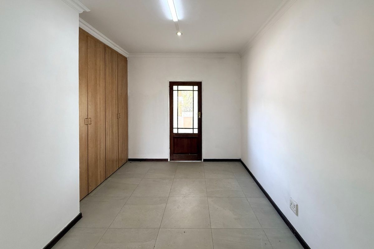 A clean, empty bedroom featuring neutral tiled flooring, built-in wooden cupboards, and a timber-framed glass door leading to the exterior.