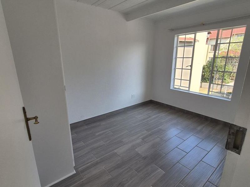 A bright, empty bedroom featuring modern wood-look tile flooring, exposed beam ceilings, and a large grid-pattern window allowing for natural light.
