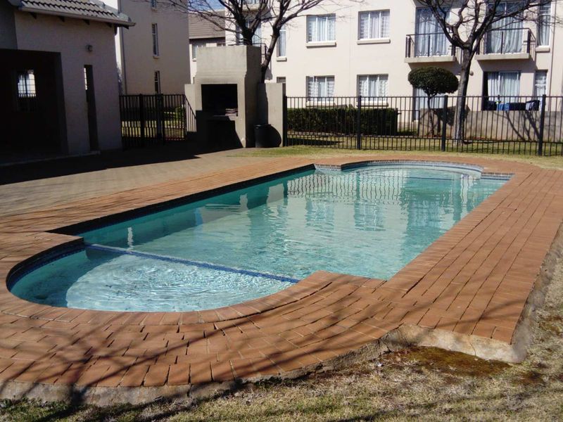 Communal swimming pool area featuring brick coping, a built-in braai, and metal perimeter fencing within an apartment complex setting.