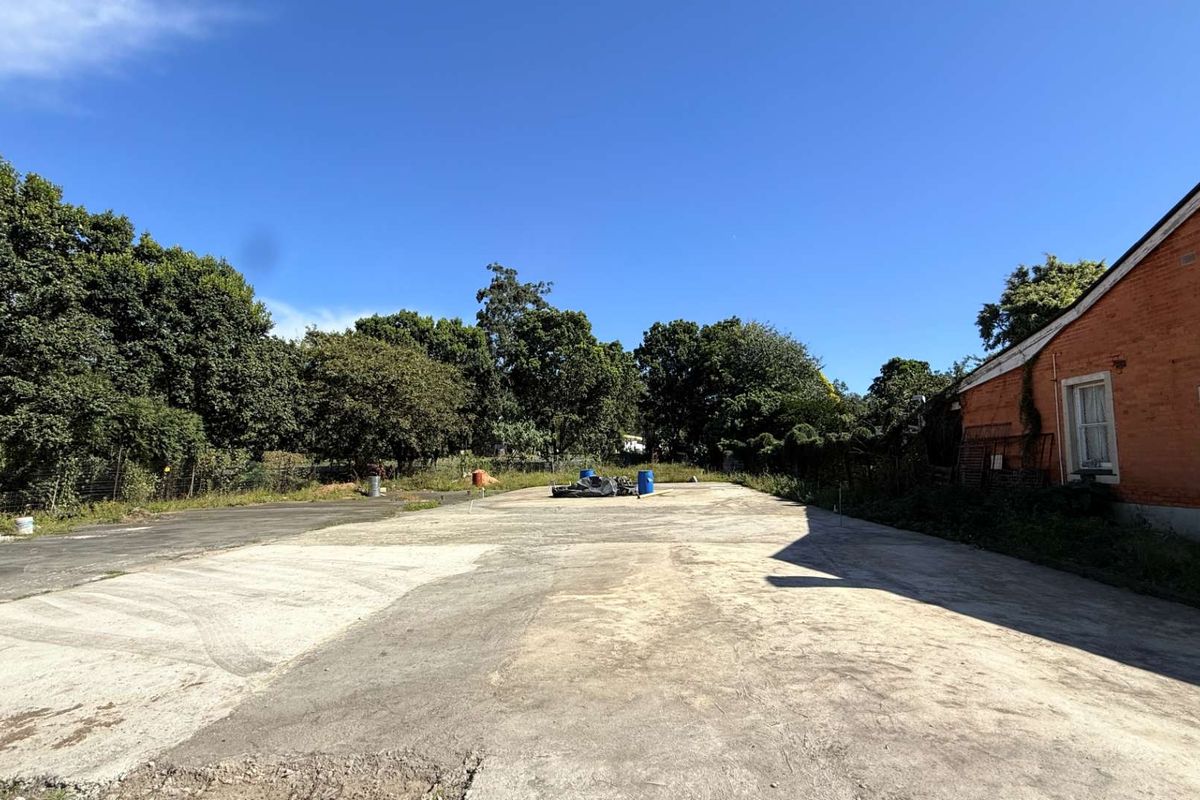 A wide view of a vacant commercial land plot featuring a large concrete paved area adjacent to an existing brick building structure.