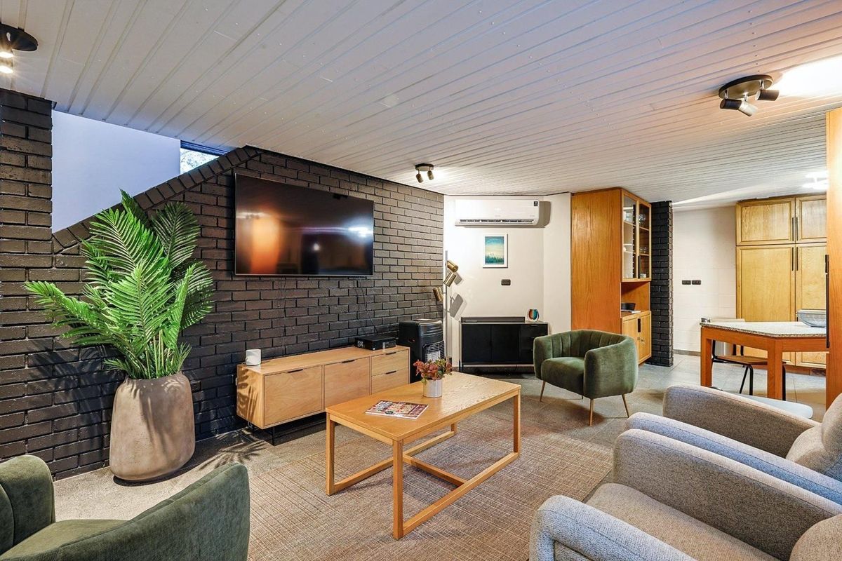 A spacious living area featuring a striking dark brick feature wall, timber-look flooring, and a split-system air conditioning unit.