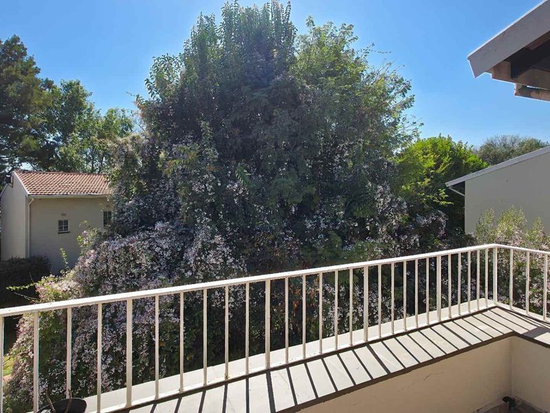A sunny balcony featuring a white metal balustrade and views of mature trees and surrounding residential buildings.