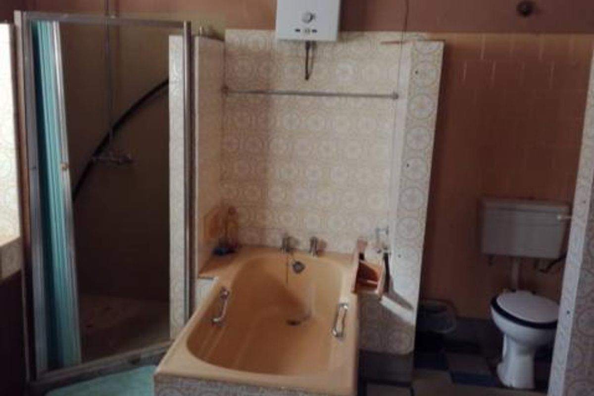 A dated bathroom featuring a built-in bathtub, separate shower enclosure, and wall-mounted gas geyser, requiring modernization of finishes and fixtures.