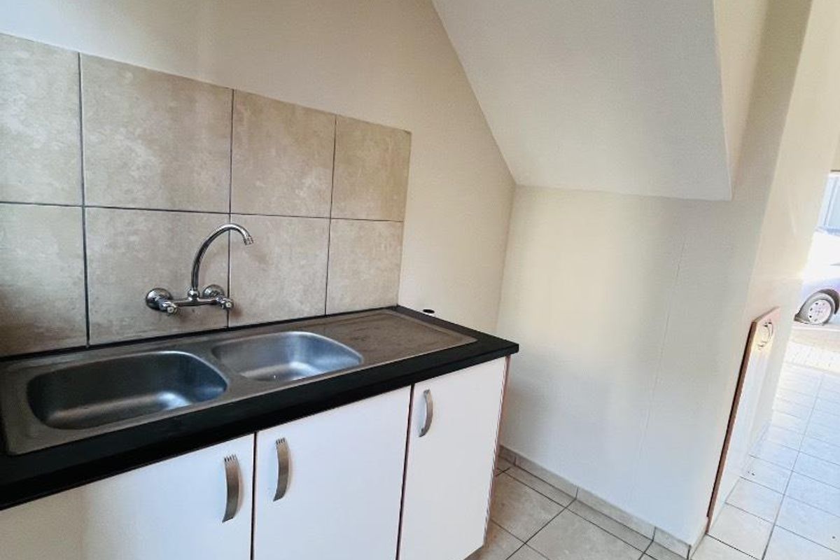 A compact kitchen area featuring a double bowl sink, tiled splashback, and white base cabinetry, situated under a sloped ceiling.