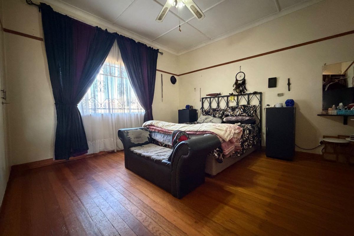 A spacious bedroom featuring original timber flooring, high ceilings with a central fan, and security bars on the window.