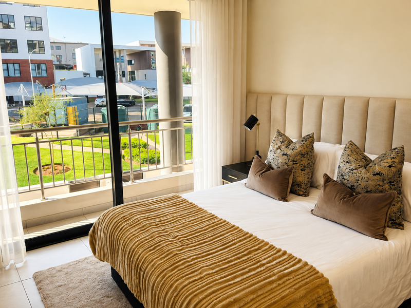 Modern bedroom featuring a neutral upholstered headboard and large sliding glass doors opening onto a private balcony with views of the complex grounds.