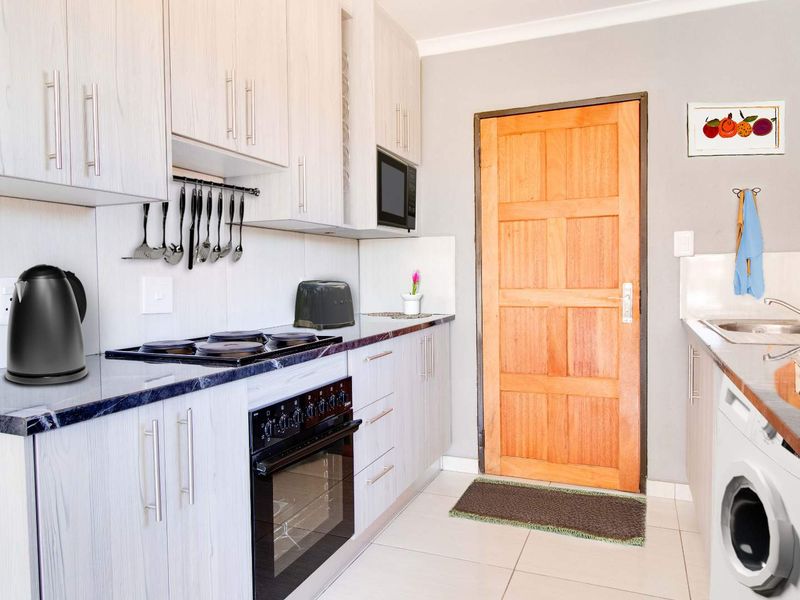 A functional kitchen featuring light wood-grain cabinetry, dark laminate countertops, built-in oven and hob, and tiled flooring with direct access to an exterior door.
