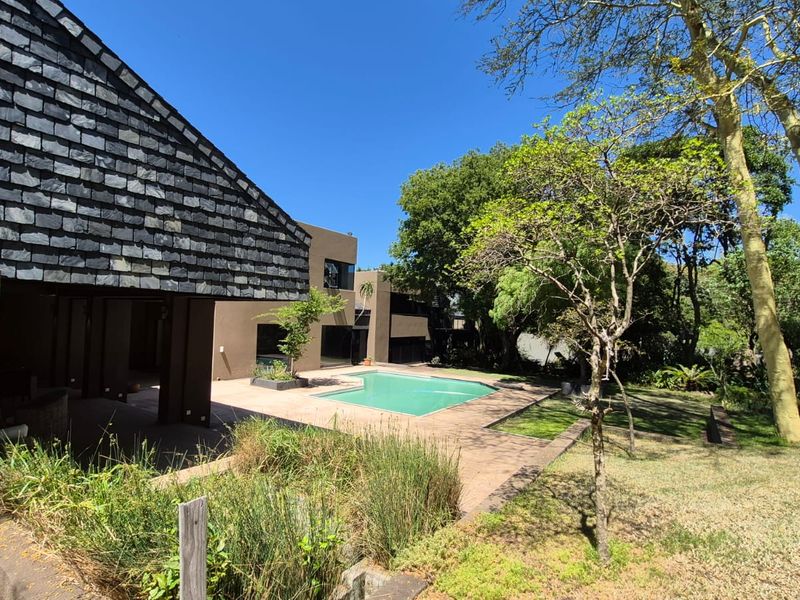 A spacious outdoor pool area featuring a paved patio, swimming pool, and distinctive slate roof architecture, set within a mature garden environment.