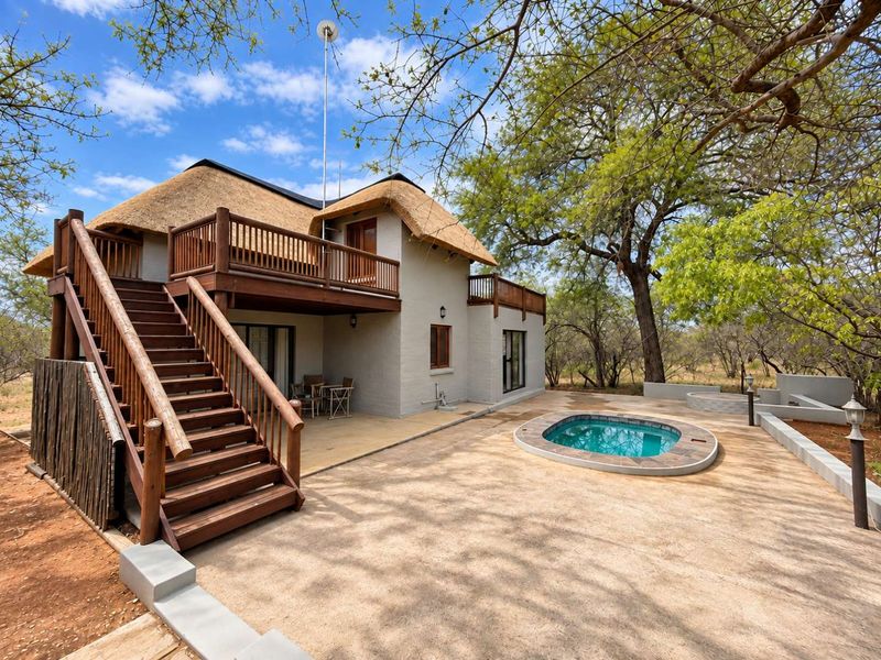 Rear view of a double-story thatched house featuring a wooden staircase, balcony, paved patio area, and a private plunge pool in a natural setting.