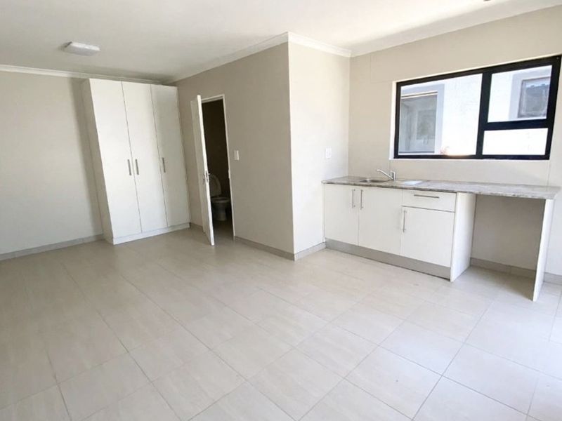 A bright, open-plan living area featuring neutral tiled flooring, built-in cupboards, and a compact kitchen kitchenette with stone countertops and a sink.