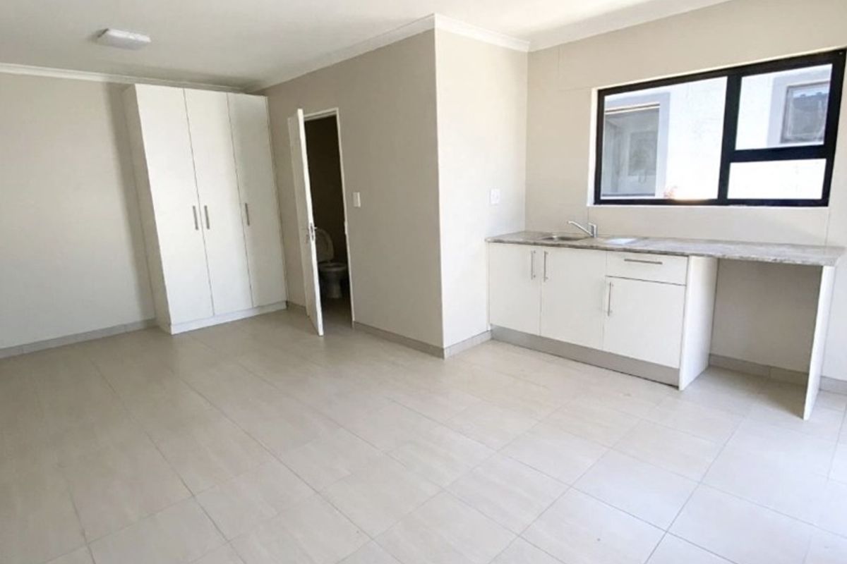 A bright, open-plan living area featuring neutral tiled flooring, built-in cupboards, and a compact kitchen kitchenette with stone countertops and a sink.