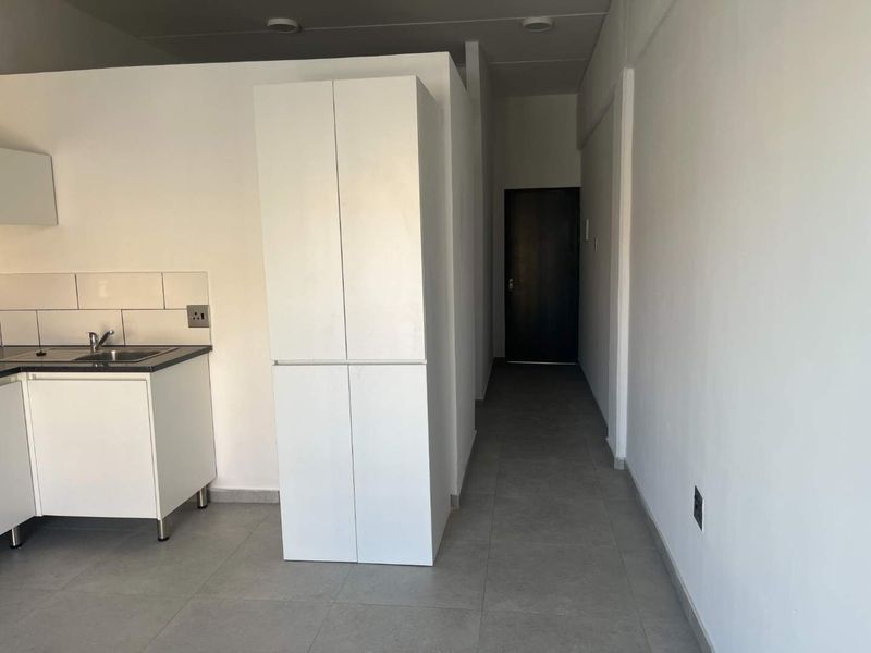 An open-plan apartment space featuring a compact kitchen area with built-in cabinetry, tiled backsplash, and neutral floor tiles, leading to an internal hallway.