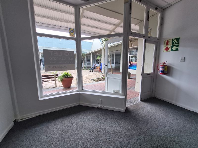 A bright, empty retail entrance space featuring large display windows, carpeted flooring, and direct access to an outdoor courtyard area.