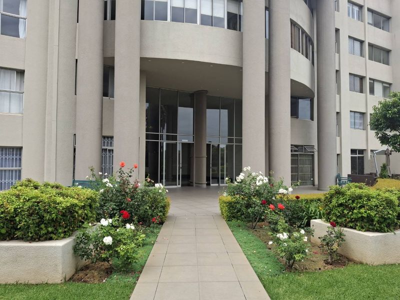 The main entrance of the apartment building features a paved walkway leading to glass doors, framed by well-maintained garden beds and concrete architectural columns.