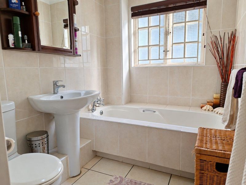 A functional bathroom featuring a pedestal basin, a built-in bathtub, and tiled finishes throughout, complemented by a window with security bars.