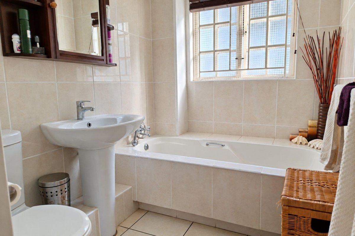 A functional bathroom featuring a pedestal basin, a built-in bathtub, and tiled finishes throughout, complemented by a window with security bars.