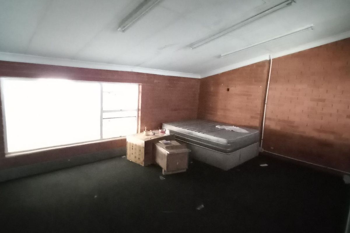 A spacious bedroom featuring exposed brick walls, large window for natural light, and fluorescent lighting, currently in need of cosmetic updates.