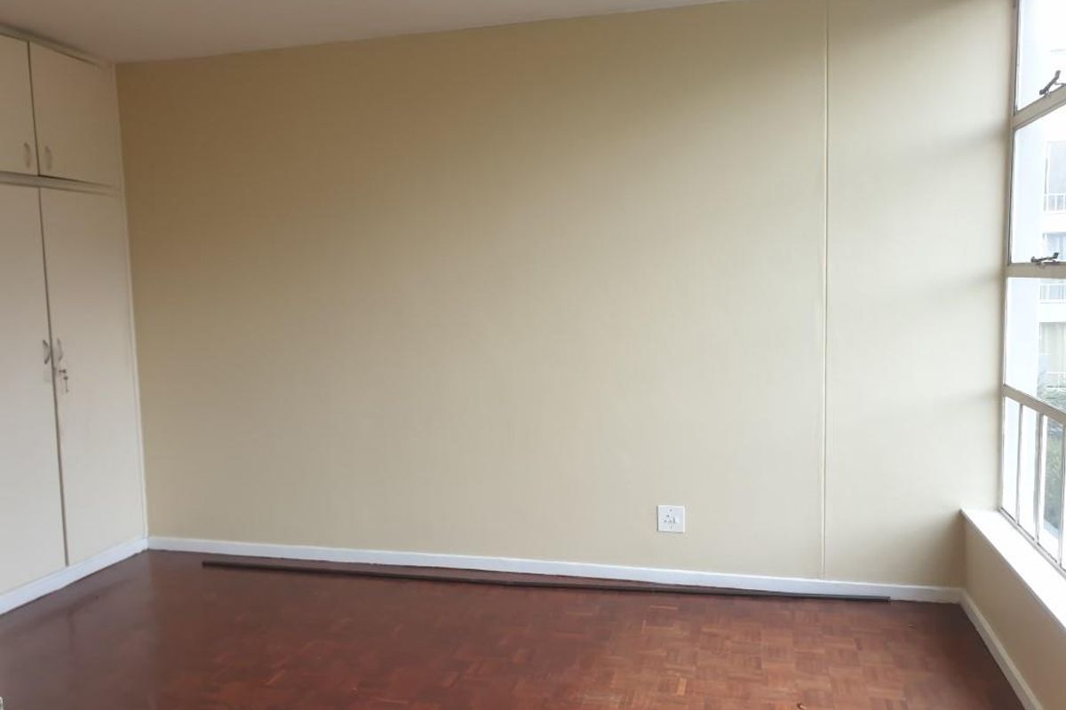 An empty bedroom featuring original parquet flooring, built-in cupboards, and a large window providing natural light.