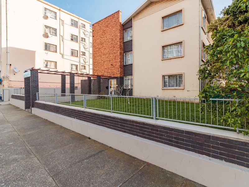Street-level view of the apartment building facade, featuring a brick boundary wall, metal fencing, and a small front garden area.