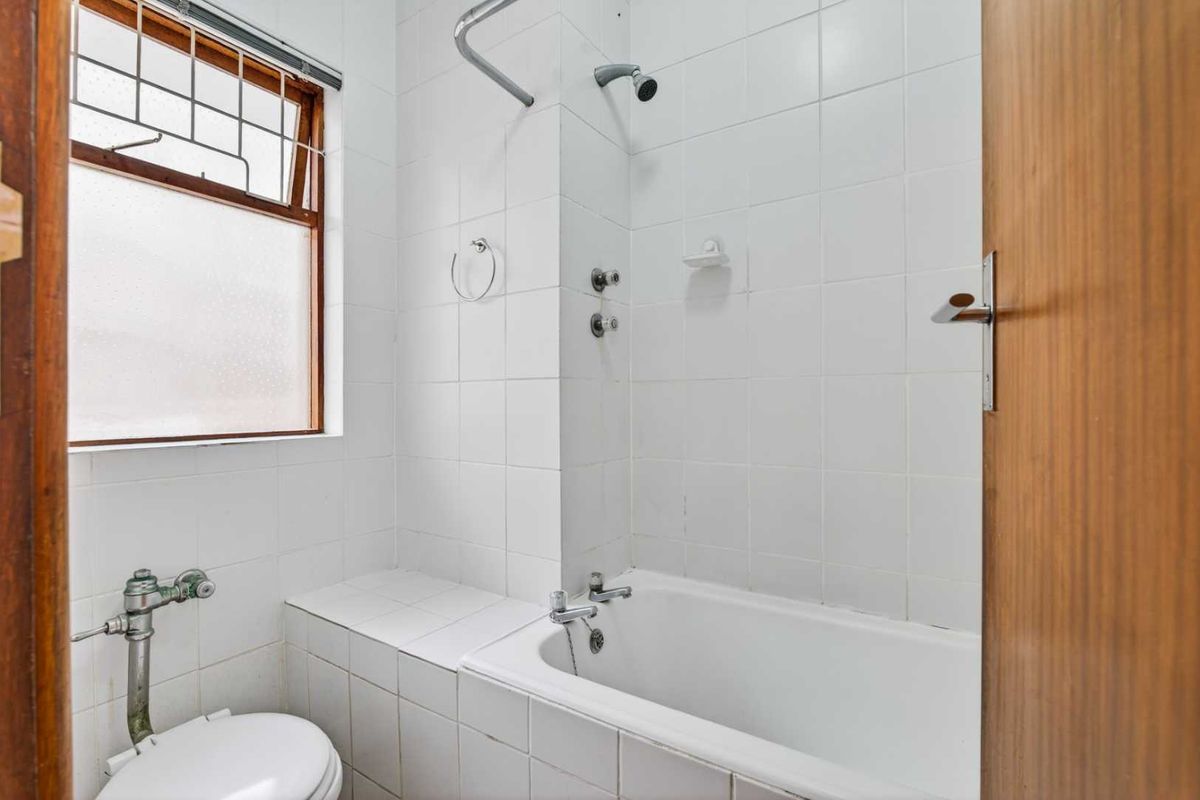 A compact bathroom featuring a built-in bathtub with shower head, white wall tiles, and a flush toilet, requiring cosmetic updates to modernize the space.