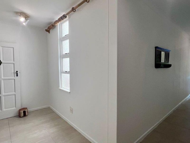 A bright, neutral hallway featuring tiled flooring, a classic panelled door, and natural light from a louvered window, leading towards an open living area.