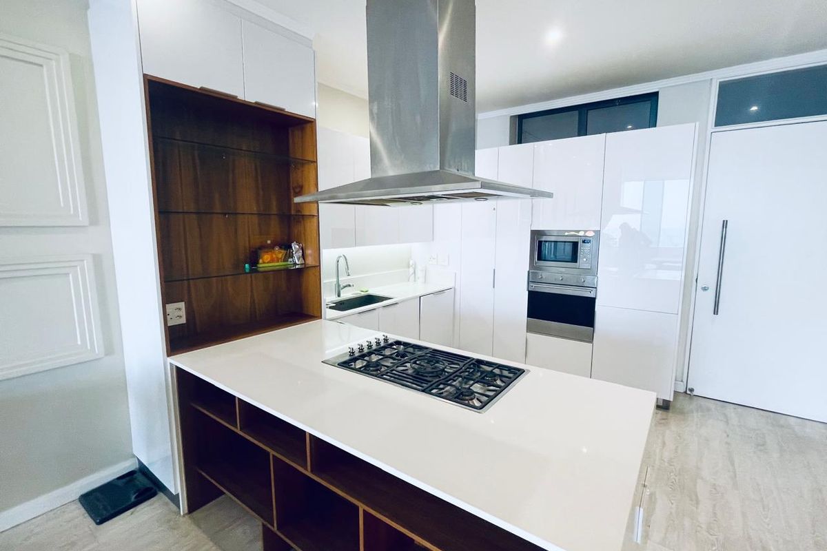 Modern kitchen featuring a central island with a gas hob, stainless steel extractor, built-in oven, microwave, and sleek white cabinetry with open shelving.