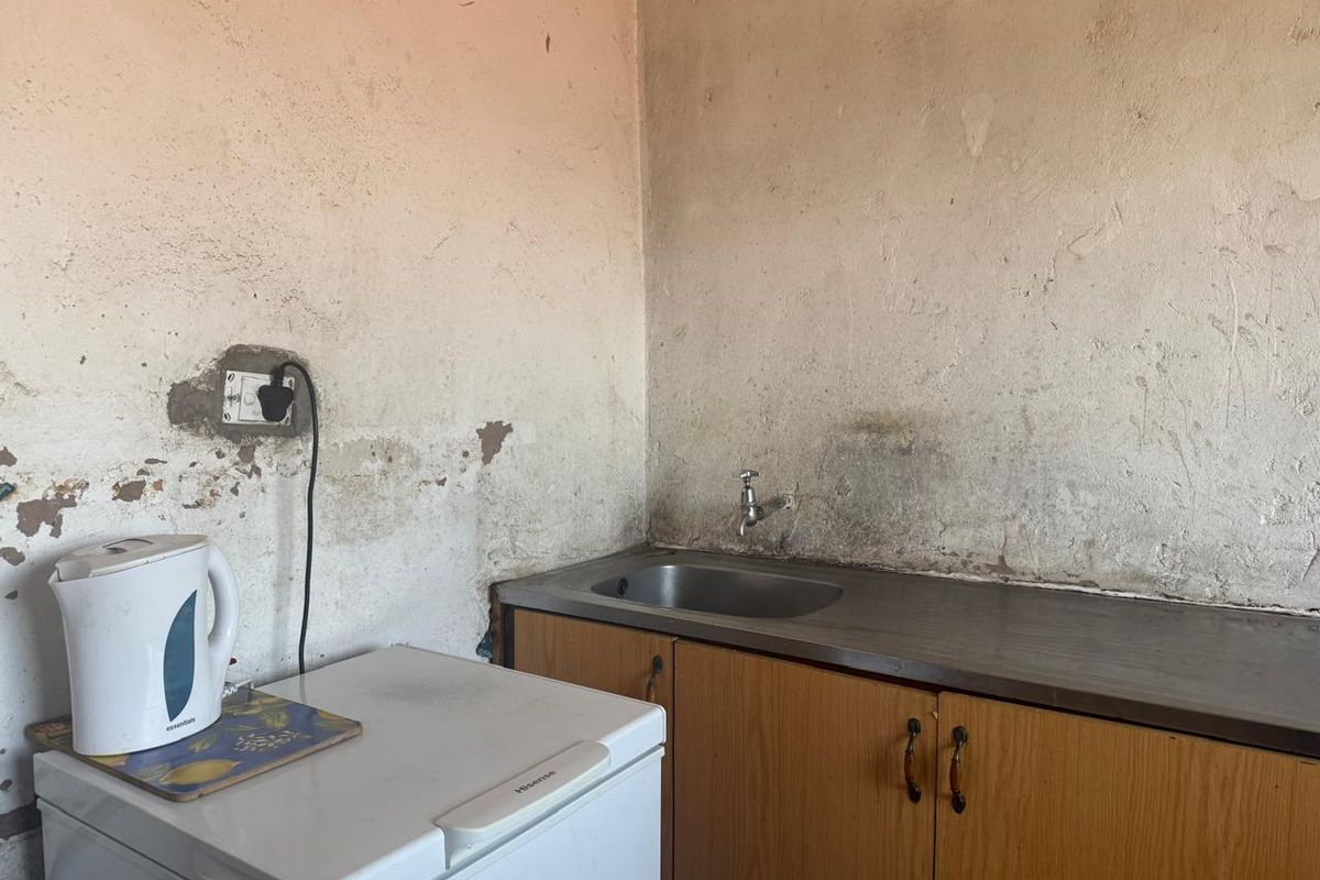 A small kitchen area featuring a stainless steel sink, built-in wooden cabinetry, and significant wall surface deterioration requiring maintenance and cosmetic repair.