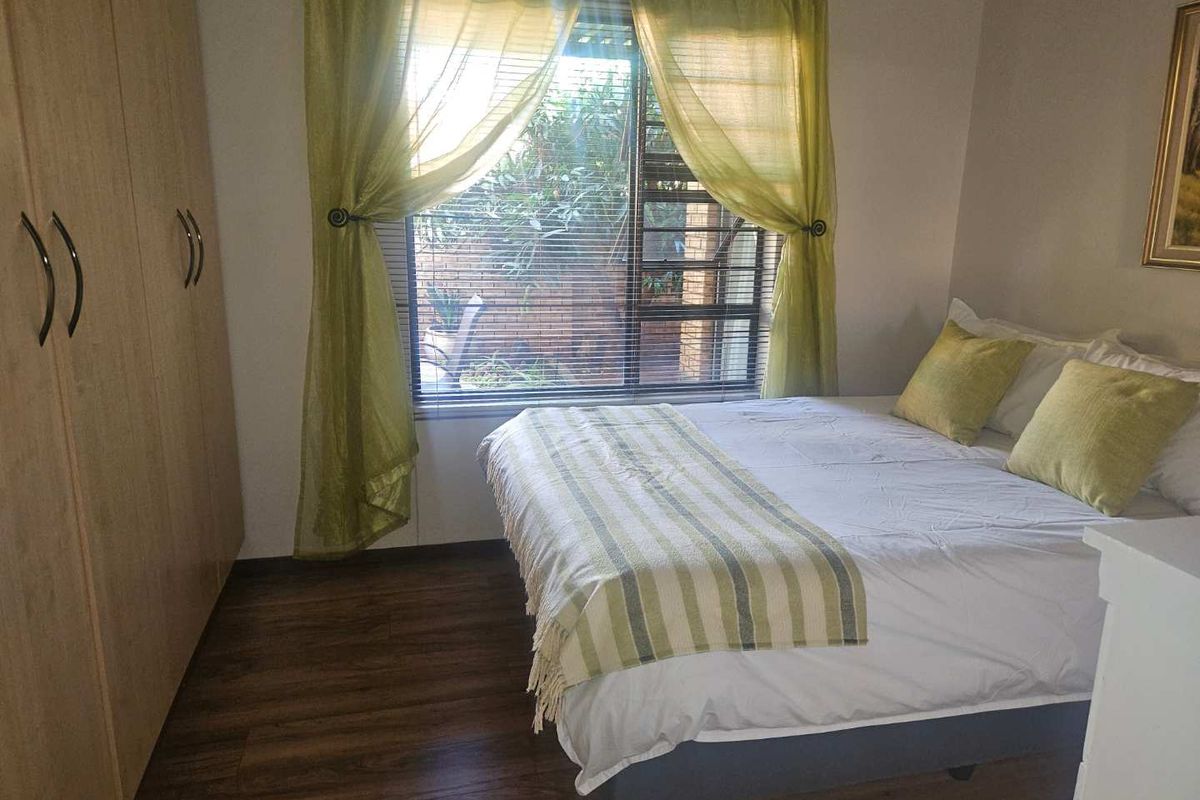 A bedroom featuring built-in wooden wardrobes, wood-look flooring, and a window with security bars overlooking a private garden area.