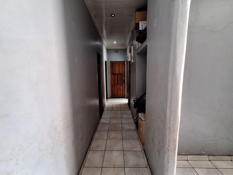 A narrow interior hallway featuring tiled flooring, recessed ceiling lighting, and a wooden door at the end.