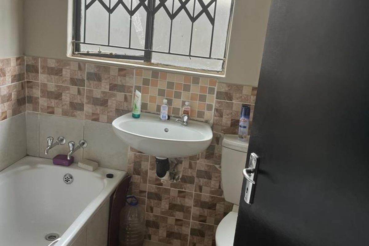 A functional bathroom featuring a standard bathtub, wall-mounted basin, and toilet, finished with neutral wall tiles and a security-grille window.