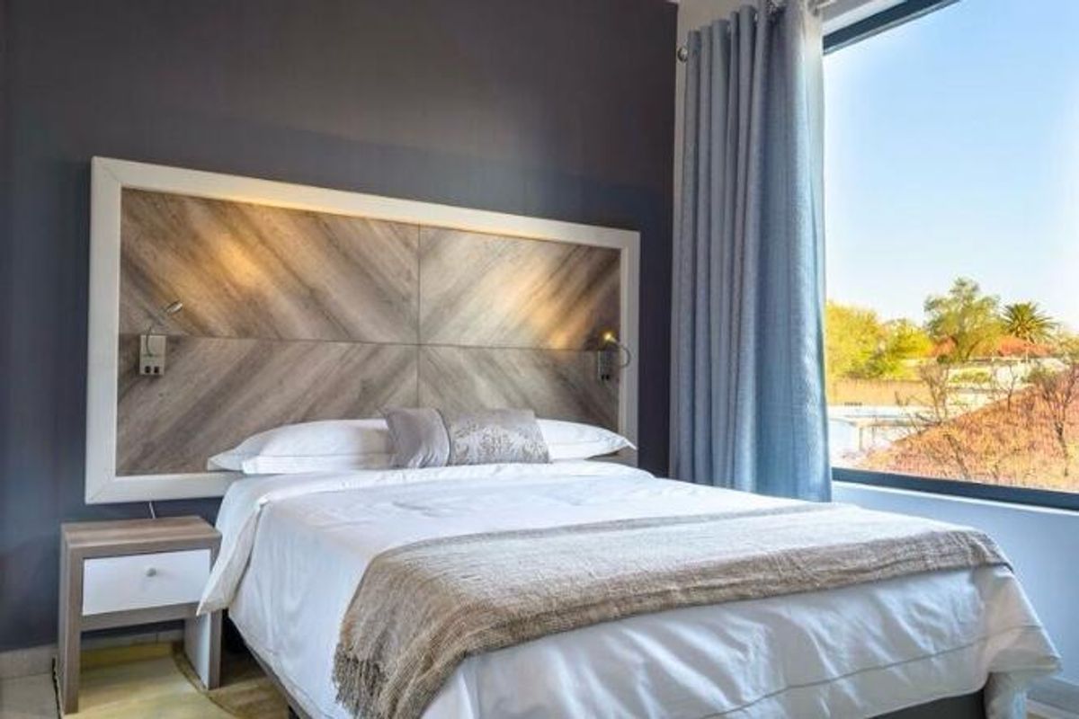 A well-maintained hotel bedroom featuring a modern wood-paneled headboard with integrated reading lights and a large window offering natural light.