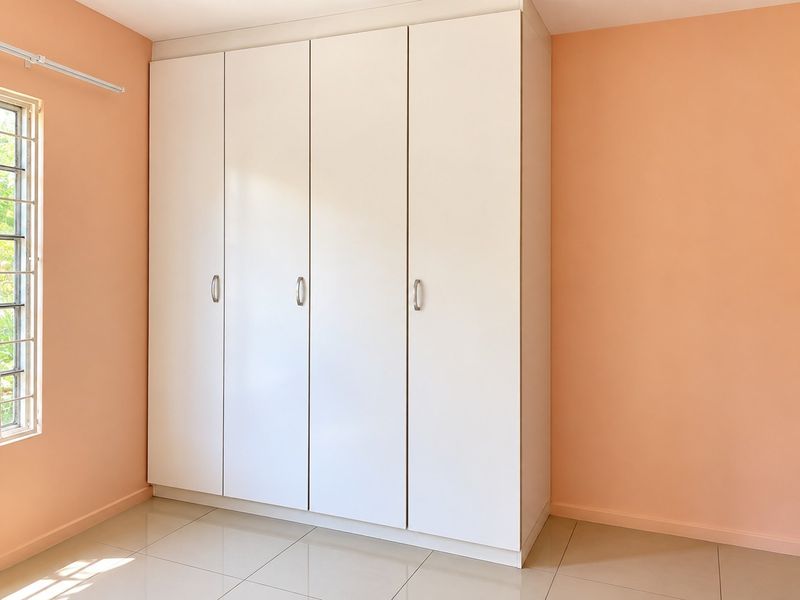 A bright, empty bedroom featuring neutral tiled flooring, peach-toned walls, and a large white built-in wardrobe with modern hardware.