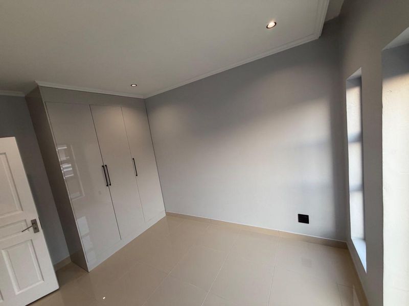 A clean, modern bedroom featuring neutral tiled flooring, built-in high-gloss cupboards, and recessed ceiling lighting.