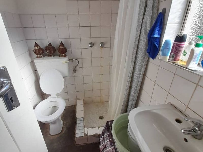 A compact bathroom featuring a shower enclosure, pedestal washbasin, and toilet, requiring renovation due to visible wear and staining on the tiled surfaces.