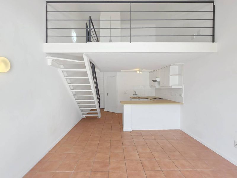 Open-plan living area featuring a mezzanine level, metal staircase, and a functional kitchen with built-in cabinetry and tiled flooring throughout.